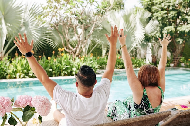 Casal maduro feliz e animado sentado em espreguiçadeiras, levantando os braços e olhando para a piscina