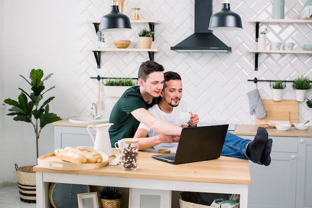 Casal masculino gay feliz abraçando enquanto estiver usando o laptop, tendo um bom tempo juntos na cozinha em casa. Conceito de casal gay feliz
