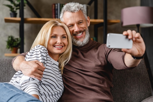 Casal moderno de meia idade sentado no sofá em casa e fazendo autorretrato no smartphone