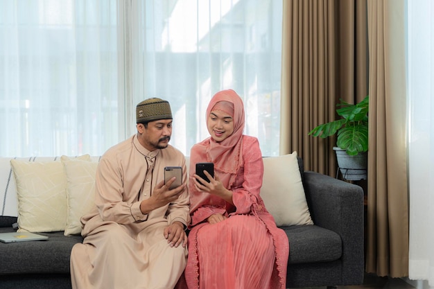 Casal muçulmano asiático fazendo uma chamada de vídeo usando um celular enquanto está sentado no sofá durante o Eid Mubarak na sala de estar
