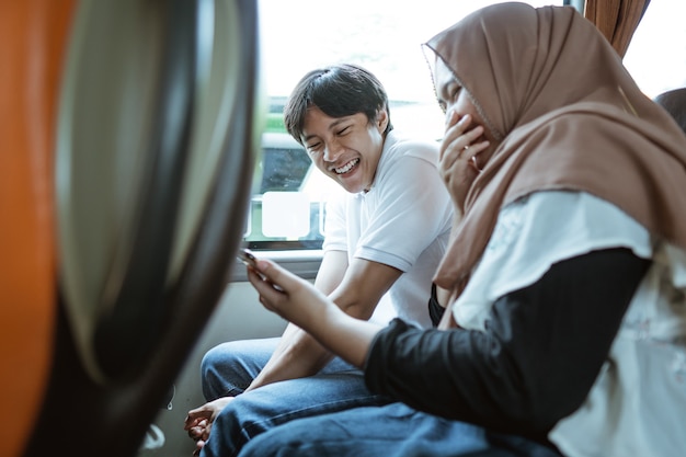 Casal muçulmano asiático ri ao ver o vídeo em seus celulares enquanto está sentado no ônibus