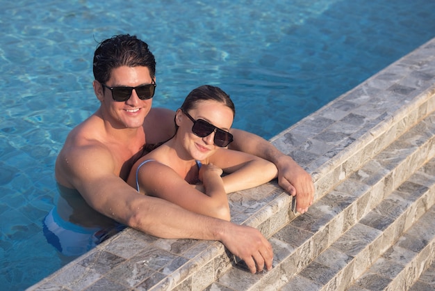 Casal muito feliz, usando óculos escuros, se divertir na piscina. Homem segurando mulher nas mãos