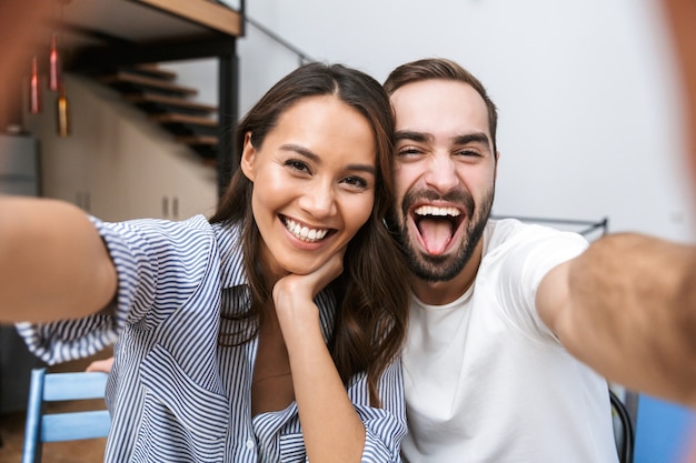 Casal multiétnico alegre tirando uma selfie enquanto está sentado na cozinha