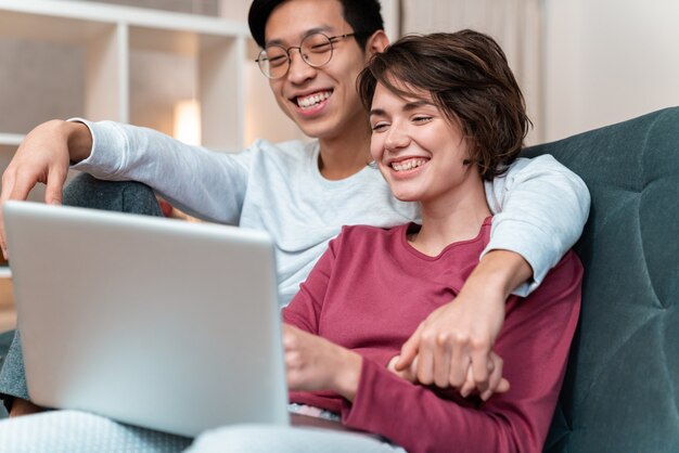 casal multinacional alegre rindo e usando laptop enquanto está sentado no sofá em casa