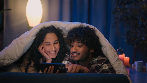 Foto casal multirracial homem e mulher à noite em casa usando telefone celular chocado espantado com notícias on-line