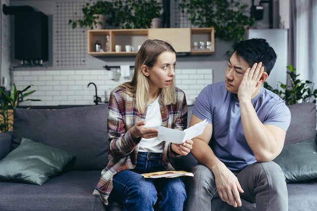 Casal multirracial homem e mulher asiáticos juntos em casa receberam uma carta com más notícias frustradas e tristes sentadas no sofá em casa