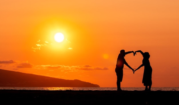 Foto casal na praia silhueta no pôr do sol amor e amizade formando coração com os braços