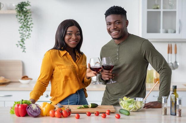 Casal negro tilintando copos com vinho na cozinha e sorrindo para a câmera