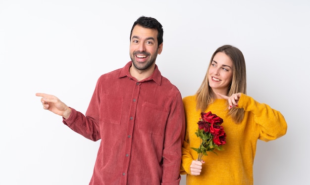 Casal no dia dos namorados, segurando flores sobre parede isolada, apontando o dedo para o lado e apresentando um produto