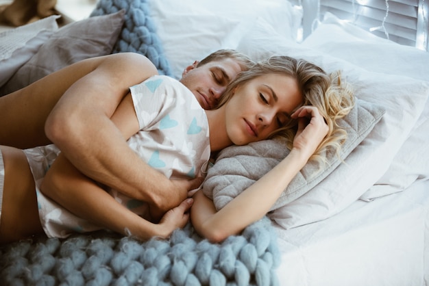 Casal no quarto em cima da cama no quarto. Uma garota loira de pijama, um cara com um torso nu. Época de Natal.