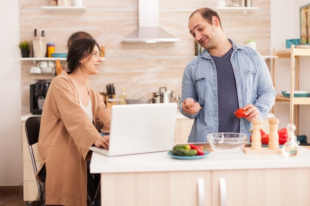 Casal olha para a receita de salada orgânica no laptop na cozinha. Homem ajudando a mulher a preparar o jantar orgânico saudável, cozinhando juntos. Relacionamento de amor romântico e alegre