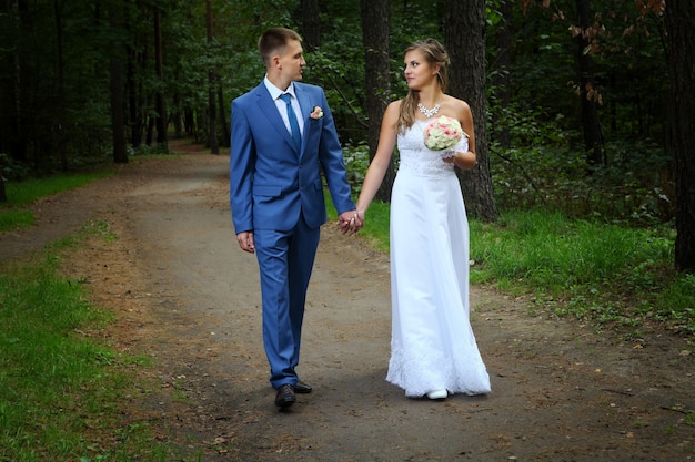 Casal recém-casado anda nos caminhos do parque de mãos dadas e olhando um para o outro.