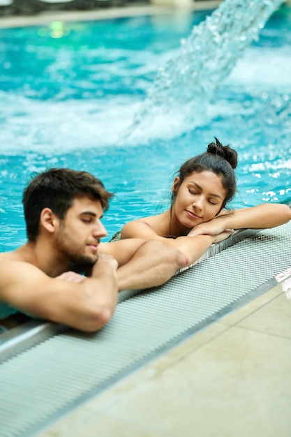 Casal relaxado, inclinando-se à beira de uma piscina e desfrutando na água com os olhos fechados no spa