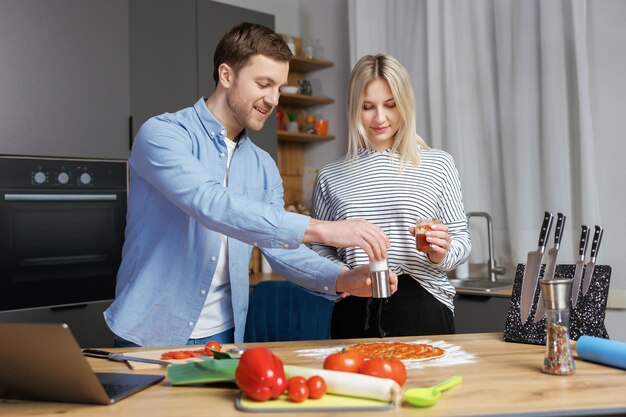 Casal romântico está cozinhando na cozinha Homem bonito e jovem atraente estão se divertindo juntos enquanto fazem salada e pizza Conceito de estilo de vida saudável