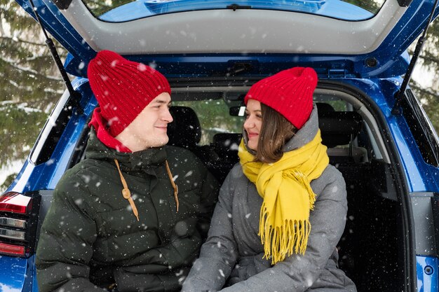 Casal romântico jovem e moderno se abraçando enquanto está sentado no porta-malas do carro sob a neve que cai