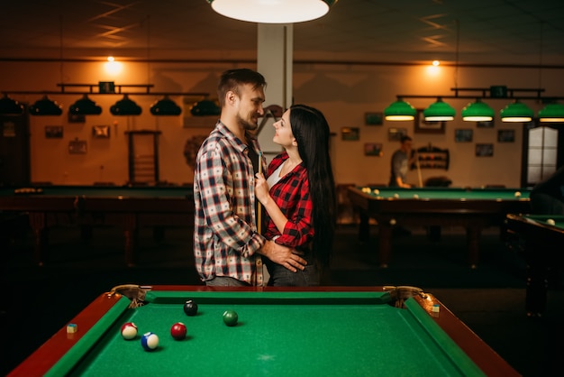 Casal se abraça à mesa com bolas coloridas na sala de bilhar. homem e mulher jogando sinuca no bar esportivo