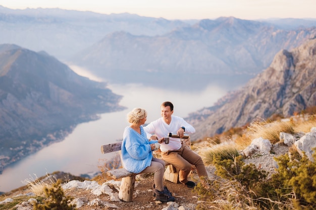 Casal se senta em um banco e bebe chá tendo como pano de fundo o Monte Lovcen e a Baía de Kotor