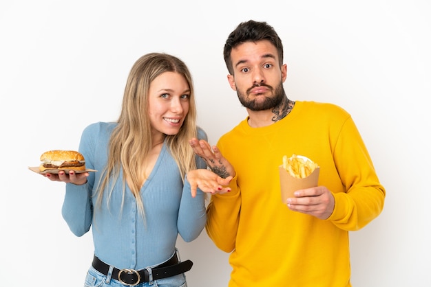 Casal segurando hambúrguer e batatas fritas sobre fundo branco isolado, tendo dúvidas enquanto levanta as mãos e os ombros