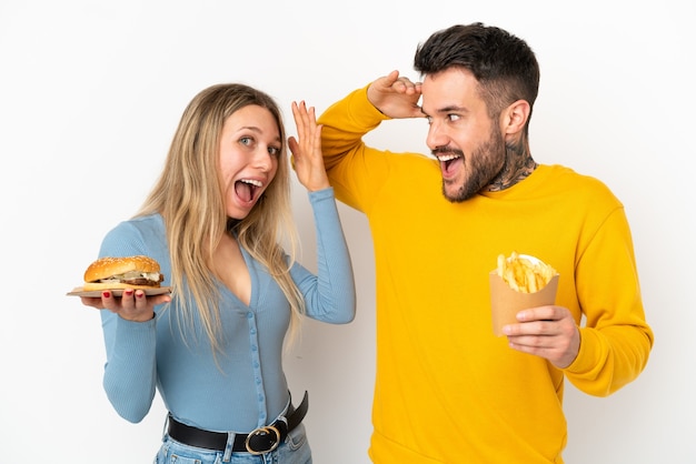 Casal segurando hambúrguer e batatas fritas sobre um fundo branco isolado com expressão facial de surpresa e choque