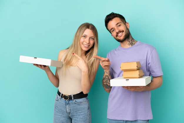 Casal segurando pizzas e hambúrgueres sobre fundo azul isolado apontando o dedo para o lado na lateral