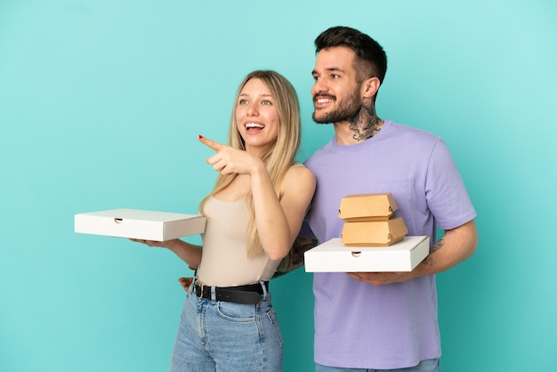 Casal segurando pizzas e hambúrgueres sobre fundo azul isolado apontando para o lado para apresentar um produto