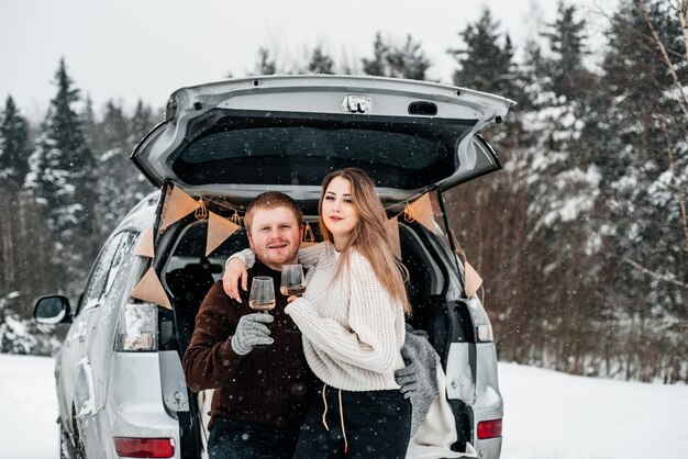 Casal segurando um copo de vinho enquanto está sentado perto do porta-malas do carro durante o inverno