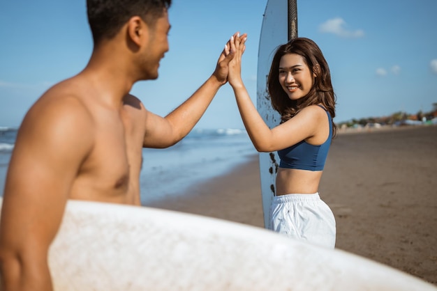 Casal segurando uma prancha de surf e dando mais cinco uns aos outros em uma praia de céu azul