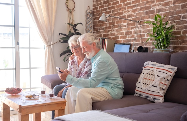 Casal sênior abraçado no sofá em casa usando telefone celular. Parede de tijolos no fundo