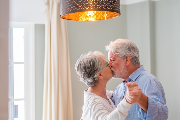 Casal sênior alegre beijando enquanto dançava em casa. Casal de idosos feliz dançando sob o lustre pendurado na sala de estar. Velho casal romântico se beijando em casa