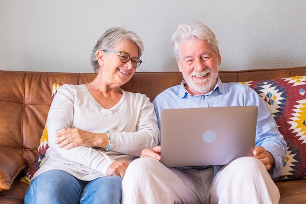 Casal sênior alegre usando laptop enquanto está sentado no sofá e sorrindo. Casal feliz de idosos relaxando enquanto navegava no laptop sentado na sala de estar. Casal idoso assistindo conteúdo de mídia usando um laptop