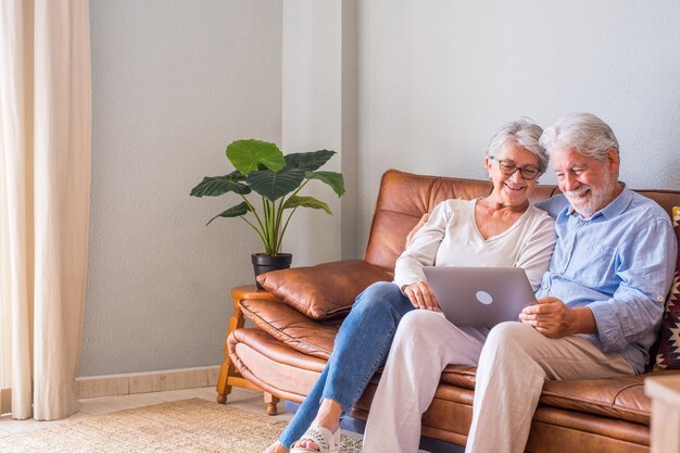 Casal sênior alegre usando laptop enquanto está sentado no sofá e sorrindo. casal feliz de idosos relaxando enquanto navegava no laptop sentado na sala de estar. casal idoso assistindo conteúdo de mídia usando um laptop