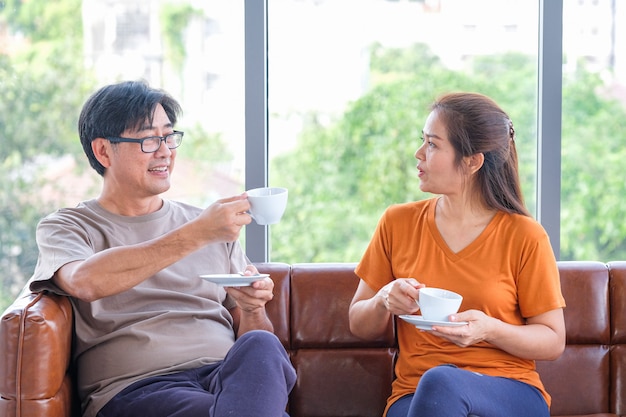 Casal sênior bebendo café, conversando e sorrindo enquanto está sentado perto da janela em casa.