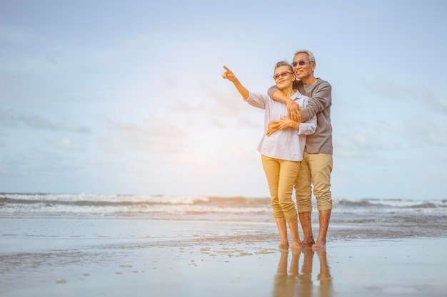 Casal sênior caminhando na praia de mãos dadas ao pôr do sol, plano de seguro de vida no conceito de aposentadoria.