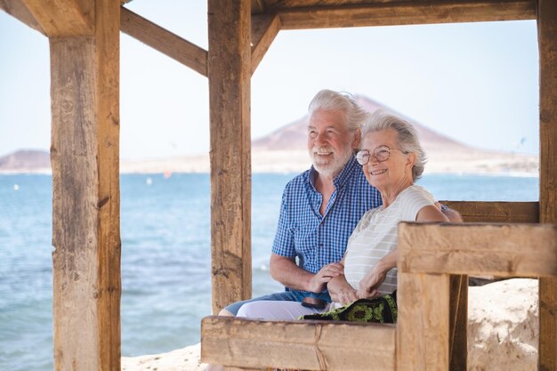 Casal sênior caucasiano feliz sentado à sombra do gazebo em frente ao mar olhando para o horizonte