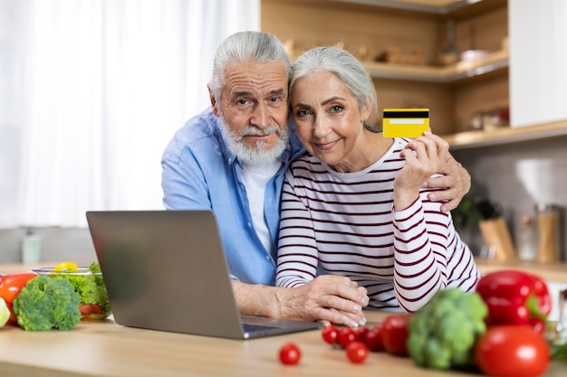 Casal sênior de conceito de comércio eletrônico segurando cartão de crédito e usando laptop na cozinha