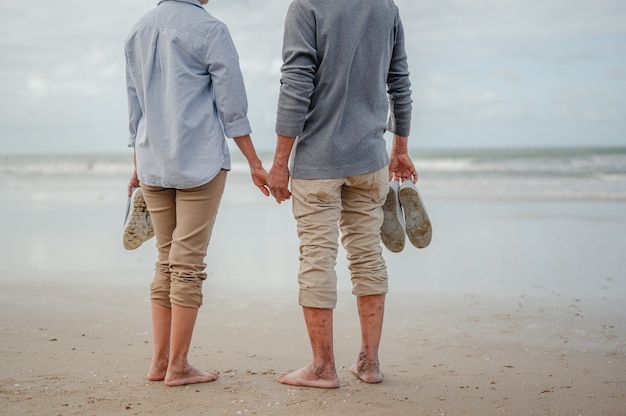 Casal sênior de mãos dadas e em pé na praia, a outra mão dos dois estava segurando um par de sapatos. Plano de seguro de vida no conceito de aposentadoria.