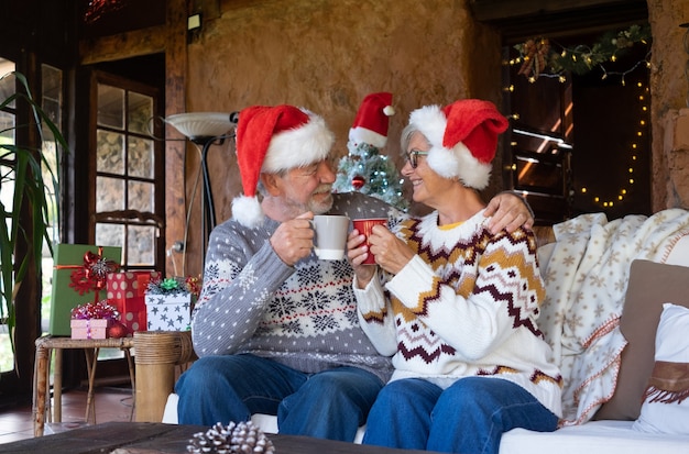 Casal sênior de sorriso no chapéu de Papai Noel, sentado no sofá em casa na época do Natal, brindando com uma xícara de chá quente. Árvore de Natal, presentes e decorações no fundo
