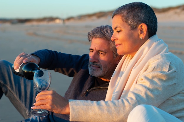 Casal sênior fazendo piquenique à beira-mar ao pôr do sol. Homem grisalho derramando vinho no copo da mulher enquanto senhora com cabelo curto sorrindo. Tiro de close-up médio. Romance, amor, conceito de relacionamento