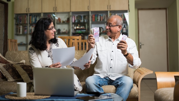 Casal sênior indiano asiático contando, fazendo finanças domésticas e verificando contas com laptop, calculadora e dinheiro também com o cofrinho, enquanto está sentado no sofá, sofá ou mesa em casa