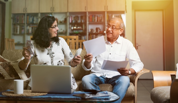 Casal sênior indiano asiático contando, fazendo finanças domésticas e verificando contas com laptop, calculadora e dinheiro também com o cofrinho, enquanto está sentado no sofá, sofá ou mesa em casa