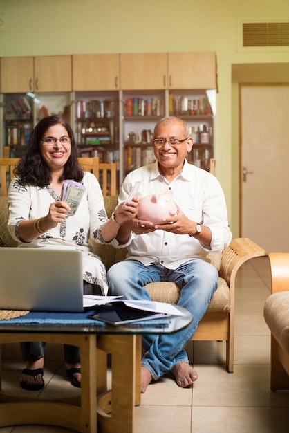 Casal sênior indiano asiático contando, fazendo finanças domésticas e verificando contas com laptop, calculadora e dinheiro também com o cofrinho, enquanto está sentado no sofá, sofá ou mesa em casa