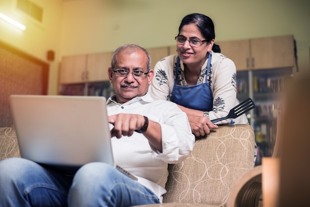 Casal sênior indiano asiático contando, fazendo finanças domésticas e verificando contas com laptop, calculadora e dinheiro também com o cofrinho, enquanto está sentado no sofá, sofá ou mesa em casa