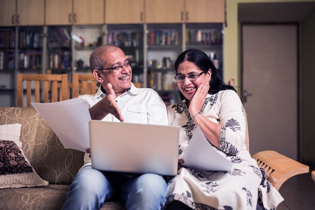 Casal sênior indiano asiático contando, fazendo finanças domésticas e verificando contas com laptop, calculadora e dinheiro também com o cofrinho, enquanto está sentado no sofá, sofá ou mesa em casa