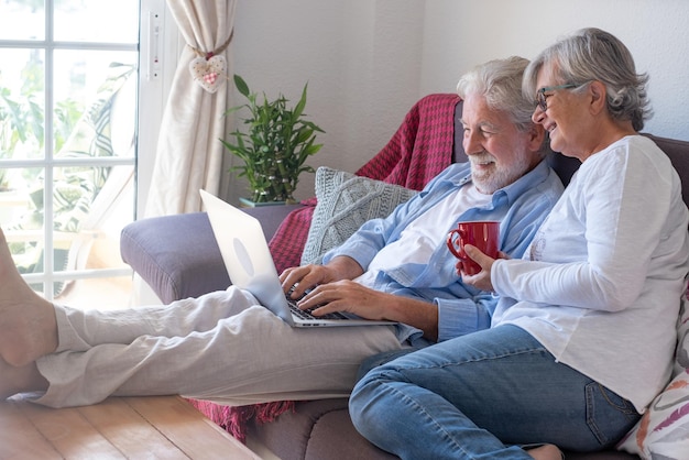 Casal sênior relaxado em casa, sentado no sofá com os pés descalços, usando o laptop. Dois idosos aposentados aproveitando o tempo livre e dispositivos tecnológicos, redes sociais