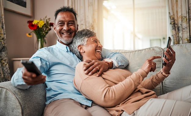 Casal sênior telefona e assiste tv no sofá de casa com um controle remoto felicidade e amor Homem e mulher maduros relaxam e riem juntos em um sofá com um smartphone ou streaming de comédia online