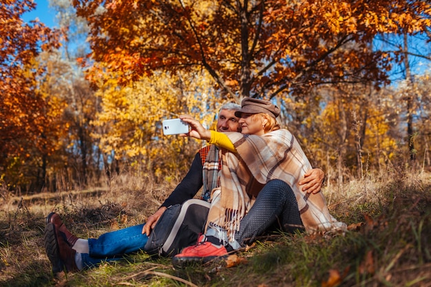 Casal sênior tomando selfie no parque outono. Homem e mulher felizes curtindo a natureza e abraçando