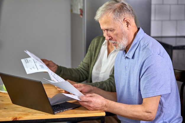 casal sênior verificando contas enquanto gerencia contas no aplicativo de home banking. homem e mulher de cabelos grisalhos casuais usando laptop enquanto olham a fatura e planejam o orçamento para economizar