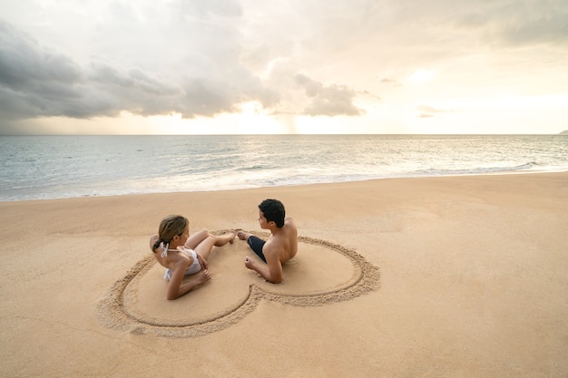 Casal sentado no coração, desenhando na praia de areia, relaxantes banhos de sol.