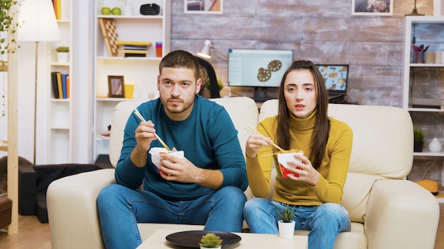 Casal sentado no sofá comendo macarrão com o pauzinho e assistindo tv.