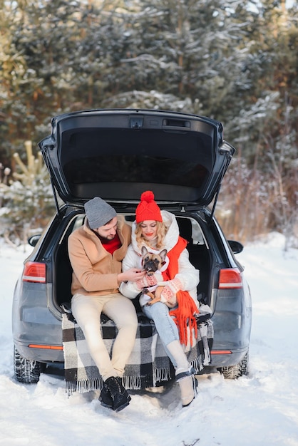 Casal sorridente com cachorro sentado no porta-malas aberto do carro SUV na floresta nevada, curtindo um ao outro nas férias de inverno ativas